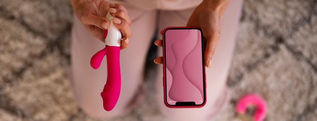 Pink silicone vibrator with white handle, curved design, held alongside smartphone for scale.
