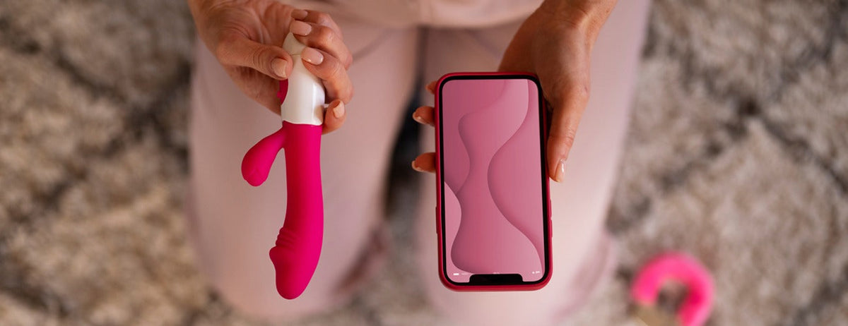Pink silicone vibrator with white handle, curved design, held alongside smartphone for scale.
