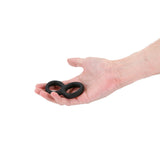 A hand holds the Renegade Twofold - Black Cock & Balls Rings, two stretchy black rubber rings linked side by side against a white background. Compatible with lubricants, they are designed to enhance pleasure.