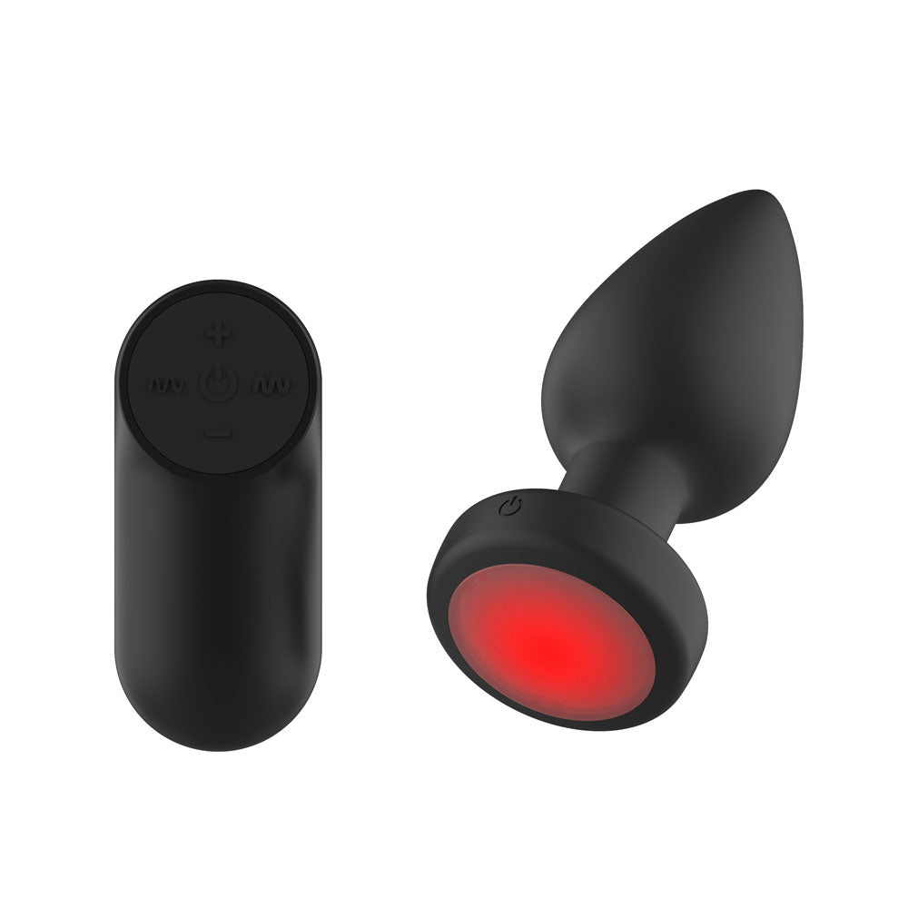 Passion Play Vibrating Butt Plug in Black with its base glowing red, oval remote at the left