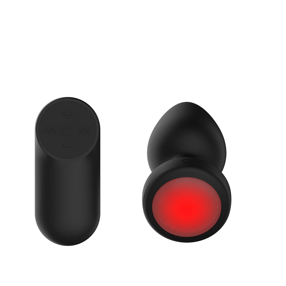 Passion Play Vibrating Butt Plug in Black tilted to show the full red LED base, remote left