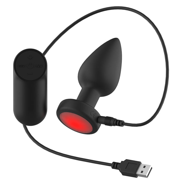 Passion Play Vibrating Butt Plug in Black on its USB charging cable, red base lit, remote left