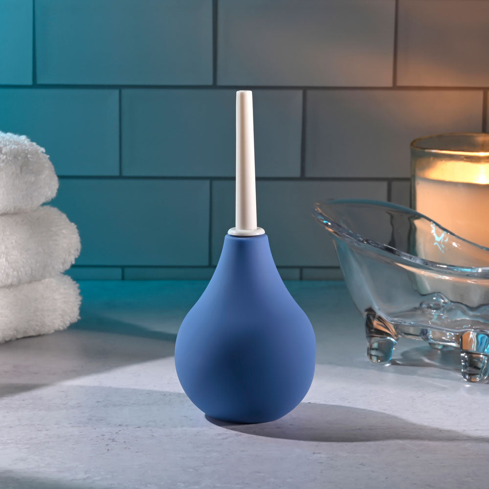 The Selopa SQUEAKY CLEAN - Blue Unisex Douche stands upright on a waterproof bathroom countertop beside neatly stacked white towels and a glass dish. A lit candle casts soft light across the tiled wall, creating an inviting ambiance for anal cleansing.