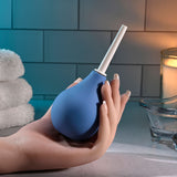 A hand holds a Selopa SQUEAKY CLEAN - Blue Unisex Douche with a white nozzle against bathroom decor featuring folded towels, a lit candle, and glass dish. Waterproof elements like softly lit tiled walls enhance the serene atmosphere. The product is phthalate and latex-free.