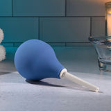 The Selopa SQUEAKY CLEAN - Blue Unisex Douche, a phthalate-free rubber bulb syringe with a white nozzle, sits on the counter against a tiled wall backdrop. A rolled towel, lit candle, and glass dish add to the serene atmosphere under soft lighting.