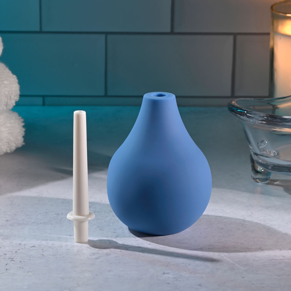 The image displays the Selopa SQUEAKY CLEAN - Blue Unisex Douche on a counter, featuring a waterproof, phthalate and latex-free rubber bulb syringe with a detachable white nozzle. A blurred candle and glass object against a tiled wall create a calm, domestic atmosphere ideal for anal cleansing.