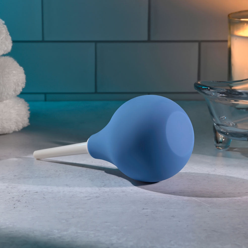 The Selopa SQUEAKY CLEAN - Blue Unisex Douche, a phthalate and latex-free rubber bulb syringe with a white nozzle, rests on a bathroom countertop. A glowing candle softly illuminates the tiled wall behind it, while rolled white towels enhance the serene ambiance of this waterproof setup for gentle cleansing.