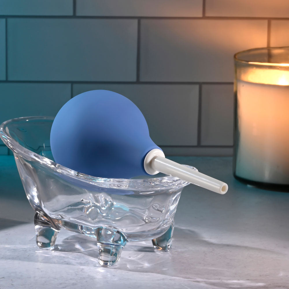 The Selopa SQUEAKY CLEAN - Blue Unisex Douche, phthalate and latex free, sits in a small bathtub-shaped clear glass dish with its white nozzle pointing outward. The waterproof dish on a smooth surface is softly illuminated by candlelight against the tile wall, creating an inviting atmosphere for cleansing.