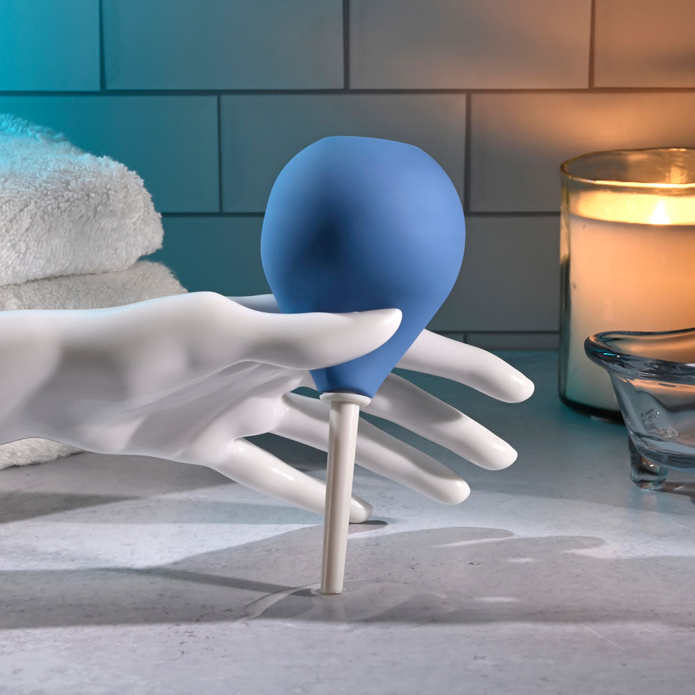 The Selopa SQUEAKY CLEAN - Blue Unisex Douche, phthalate and latex-free, is gripped by a white mannequin hand on a bathroom counter. Behind it are stacked white towels, a lit candle, and a clear dish against white tiles. Soft shadows add to the serene ambiance.