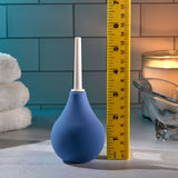 A Selopa SQUEAKY CLEAN - Blue Unisex Douche lies beside a yellow measuring tape on a bathroom counter. The background features folded white towels, a lit candle, a glass dish, and a white tile wall, all under soft ambient lighting for a serene atmosphere.