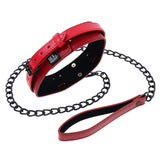 The S&M Exs & Ohs Red Neck Restraint features a red faux leather collar with an adjustable buckle and is attached to a matte black chain leash. It has a matching red leather handle, metal snap closure, silver round studs, and a small label reading SEX & MISCHIEF.