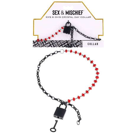 The S&M Exs & Ohs Crystal Day Collar features ruby red crystals on a matte black chain with a lock and key. It includes elegant packaging: a white card bordered in gold with sophisticated black text and decorative elements.