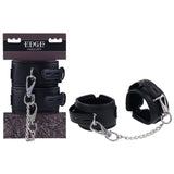 The EDGE Handcuffs - Black Restraints feature metal buckles and a chain for secure restraint, with adjustable buckle closures. Displayed both in and out of their packaging, the box showcases EDGE and HANDCUFFS with a geometric pattern.