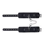 The EDGE Handcuffs - Black Restraints have black leather with an adjustable buckle, secure constraint design, padded interiors for comfort, and metal rivets for durability. A short silver chain with carabiners connects the cuffs.
