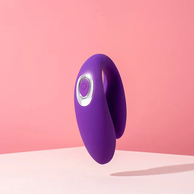 A purple C-shaped couples vibrator balanced on a soft pink backdrop