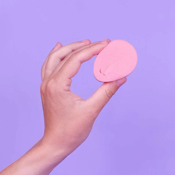 Pink teardrop-shaped makeup sponge in soft foam, held in hand against purple background.
