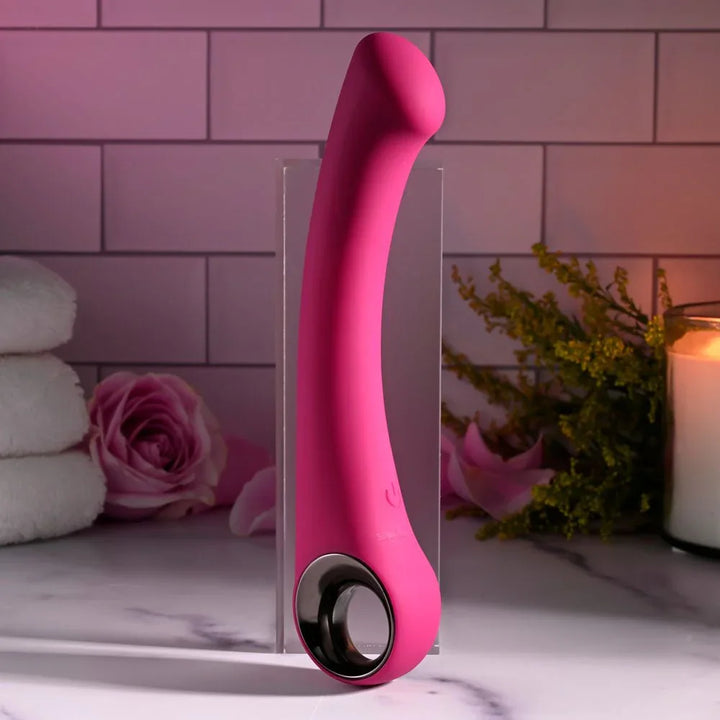Evolved Pleasure Curve in Pink upright against a clear block, roses and folded towels behind