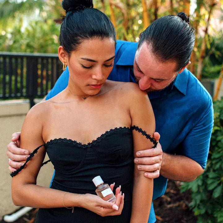 Man kissing a woman's bare shoulder while she holds the After Dark perfume bottle