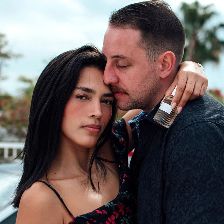 Couple embracing outdoors, the woman holding an Eye Of Love After Dark bottle to camera