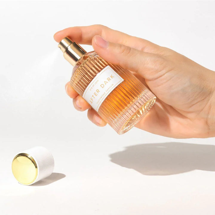 A hand spraying the Eye Of Love After Dark 50ml bottle, gold cap lying on the surface