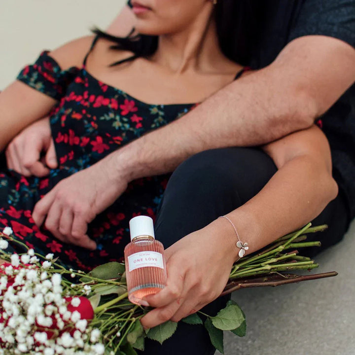 Seated woman in a floral dress holding a One Love perfume bottle, roses in her lap