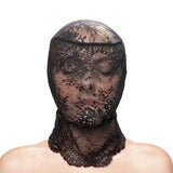 With closed eyes, a person wears the Fetish & Fashion - Lace Hood - Black over their head and shoulders. The form-fitting black lace, with intricate floral patterns, partially obscures their face against a white background that highlights the fabrics details.