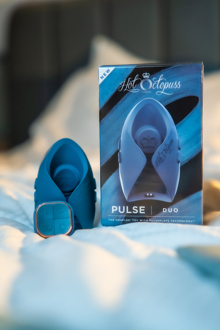 Hot Octopuss Pulse Duo in blue with its remote, beside the retail box on a bed