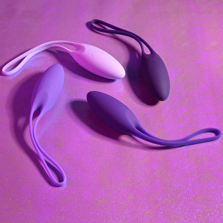 Purple Put In Work kegel balls scattered on a pink surface, silicone cords looped