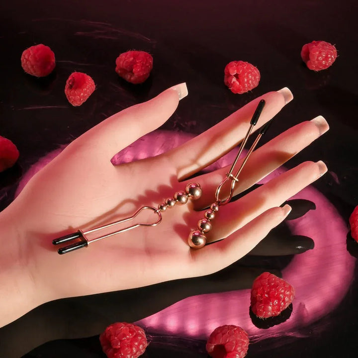 A hand holding one rose gold beaded nipple clamp, bead tail hanging