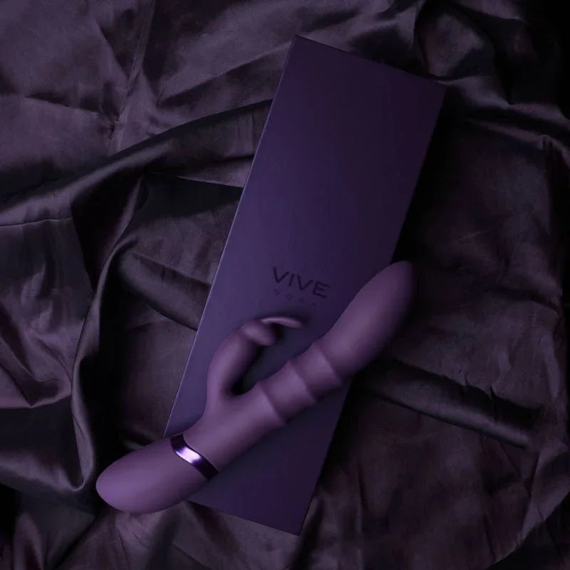 VIVE Sora Rabbit Vibrator in Purple, lifestyle shot lying on black satin beside its box